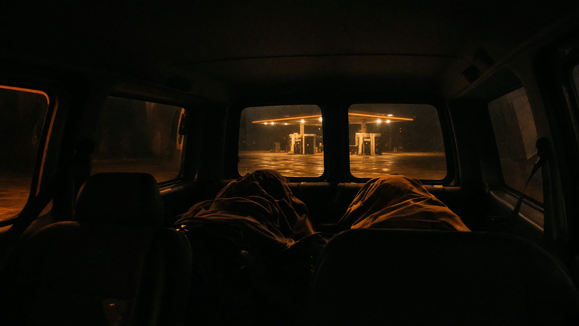 Four people asleep under coats in the back of a van at a service station at night